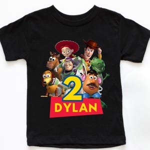 May include: Black short-sleeved t-shirt featuring a colorful graphic of characters from the animated film Toy Story. The number "2" is prominently displayed, along with the name "DYLAN" in white letters on a red background.