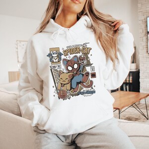 Puede incluir: Sudadera blanca con un estampado gr&aacute;fico de un gato de dibujos animados vestido de Spider-Man. El gr&aacute;fico incluye el texto "The Ameowzing Spider-Cat" y "The Origin of Peter Purrke".