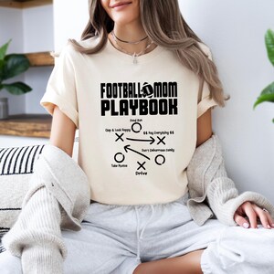 May include: A white t-shirt with a black graphic that says "Football Mom Playbook" and includes a football and a play diagram. The diagram includes the text "Clap & Look Happy", "Food Run", "$$ Pay Everything $$", "Don't Embarrass Family", "Take Photos", and "Drive".