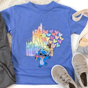 Puede incluir: Una camiseta azul con un personaje de dibujos animados sosteniendo un helado y globos. El gr&aacute;fico incluye un castillo colorido y globos en forma de coraz&oacute;n. La camiseta se combina con pantalones grises y zapatos beige.