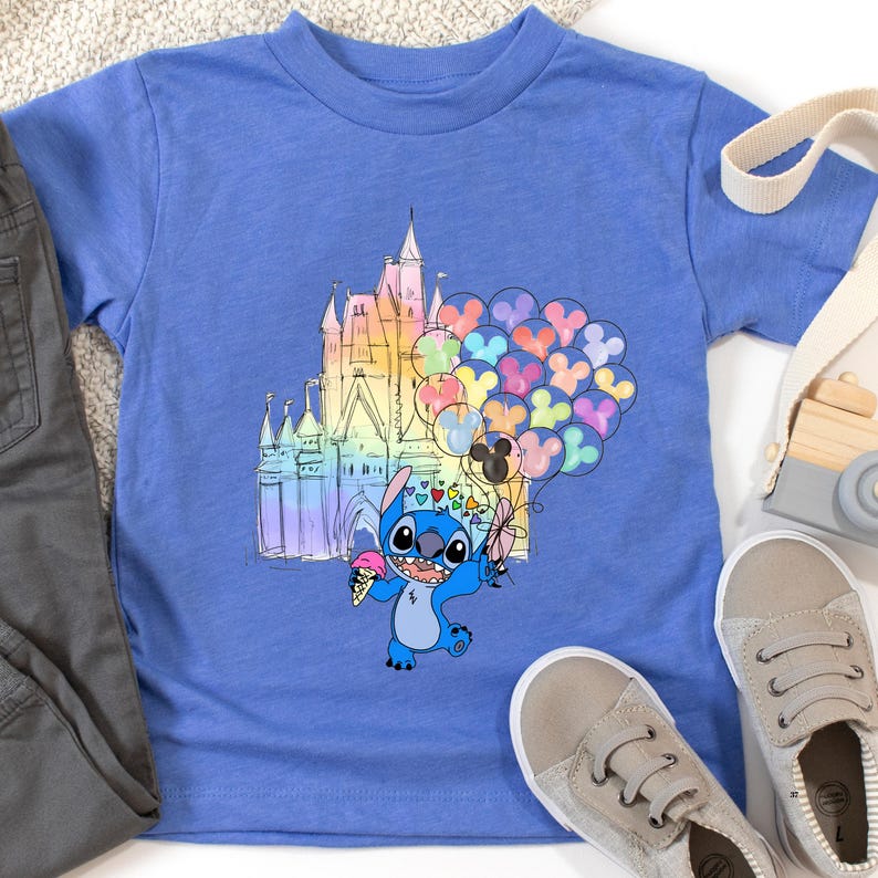 Puede incluir: Una camiseta azul con un personaje de dibujos animados sosteniendo un helado y globos. El gr&aacute;fico incluye un castillo colorido y globos en forma de coraz&oacute;n. La camiseta se combina con pantalones grises y zapatos beige.