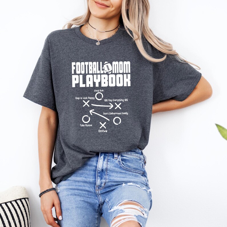 May include: A gray t-shirt with the text "Football Mom Playbook" and a football graphic. The shirt features a diagram with the text "Dap & Look Happy", "Food Run", "$$ Pay Everything $$", "Don't Embarrass Family", "Take Photos", and "Drive".