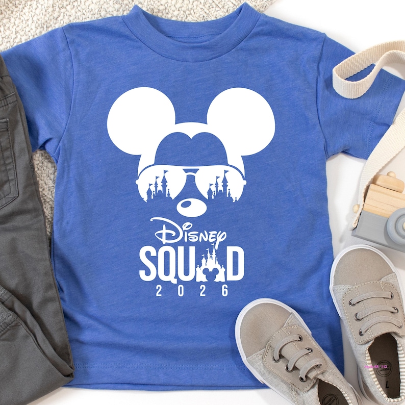 May include: A blue t-shirt featuring a white Mickey Mouse head wearing sunglasses with a castle design. The text "Disney Squad 2026" is printed below the graphic. Gray pants and shoes are visible.