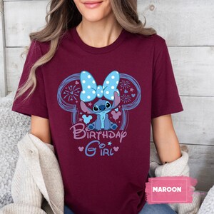 May include: Maroon t-shirt featuring a cartoon Stitch character with a blue bow and the words "Birthday Girl." The design includes a blue outline of Mickey Mouse ears and fireworks. The shirt is a solid maroon color.