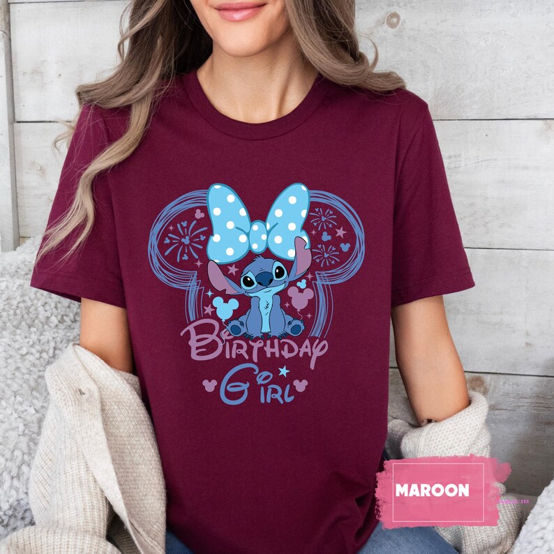 May include: Maroon t-shirt featuring a cartoon Stitch character with a blue bow and the words "Birthday Girl." The design includes a blue outline of Mickey Mouse ears and fireworks. The shirt is a solid maroon color.