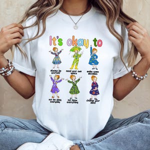 May include: White t-shirt with the text "It's okay to" above colorful illustrations of women in dresses. The illustrations are accompanied by phrases like "always be yourself" and "make some mistakes."