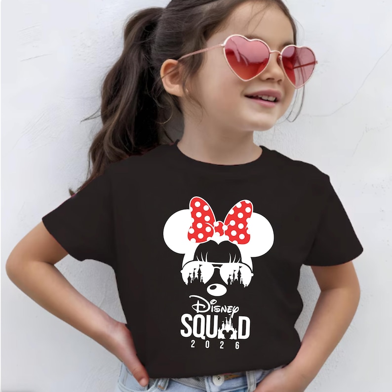 May include: Black t-shirt featuring a Minnie Mouse graphic with a red polka dot bow, sunglasses, and the text "Disney Squad 2026". The graphic is white. The girl is wearing heart-shaped sunglasses.