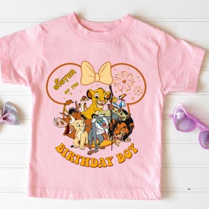 May include: Pink short-sleeved t-shirt featuring a graphic of characters from The Lion King, including Simba, Timon, and Pumbaa. The text "Sister of the Birthday Boy" is printed in a circular design around the characters. A yellow bow and fireworks complete the design.