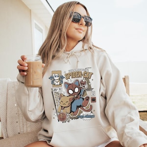 Puede incluir: Una sudadera con capucha blanca con un estampado gr&aacute;fico de un gato de dibujos animados vestido de Spider-Man. El gr&aacute;fico incluye el texto "The Ameowzing Spider-Cat" y "The Origin of Peter Purrker".