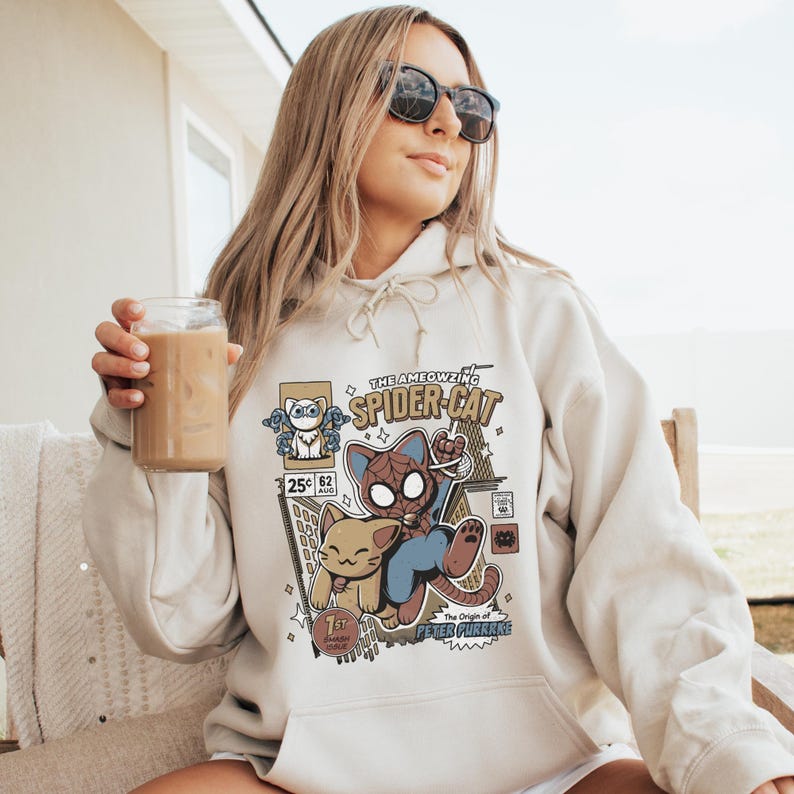 Puede incluir: Una sudadera con capucha blanca con un estampado gr&aacute;fico de un gato de dibujos animados vestido de Spider-Man. El gr&aacute;fico incluye el texto "The Ameowzing Spider-Cat" y "The Origin of Peter Purrker".