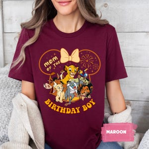 May include: Maroon t-shirt featuring a graphic of characters from The Lion King, with the text "Mom of the Birthday Boy." The design includes Simba, Timon, Pumbaa, and other characters. A yellow bow and fireworks complete the design.