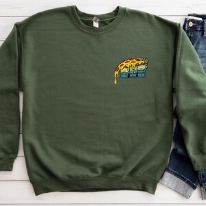 May include: Dark green sweatshirt with a small graphic of three cartoon characters under a slice of pizza. The pizza has yellow, red, and brown toppings with melted cheese dripping down. The sweatshirt is a solid color and has a crew neck.