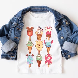 May include: White t-shirt featuring colorful cartoon ice cream designs. The ice cream popsicles have various character faces and patterns. A denim jacket is draped over the shirt.