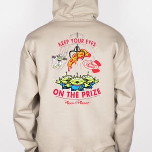 May include: Beige hoodie featuring a graphic from the movie Toy Story. The design includes the text "KEEP YOUR EYES ON THE PRIZE" with images of the Pizza Planet aliens, Buzz Lightyear, and a pizza slice.