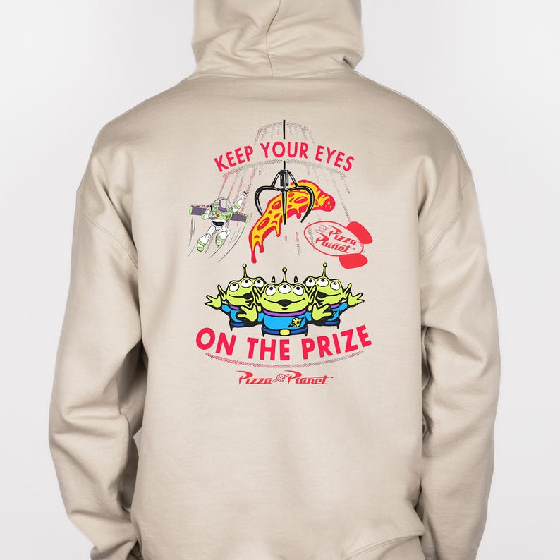 May include: Beige hoodie featuring a graphic from the movie Toy Story. The design includes the text "KEEP YOUR EYES ON THE PRIZE" with images of the Pizza Planet aliens, Buzz Lightyear, and a pizza slice.