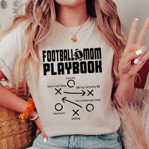 May include: A beige t-shirt with the text "FOOTBALL MOM PLAYBOOK" in black. The shirt features a football play diagram with instructions like "Clap & Look Happy", "Food Run", "$$ Pay Everything $$", "Don't Embarrass Family", "Take Photos", and "Drive".