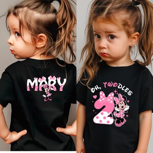 K&ouml;nnte beinhalten: Schwarze T-Shirts mit Minnie-Maus-Designs. Ein Shirt tr&auml;gt die Aufschrift "MARY" mit einer Minnie-Maus-Grafik. Das andere Shirt tr&auml;gt die Aufschrift "OH, TWOODLES" mit der Zahl 2 und Minnie Maus. Beide Shirts haben rosa Akzente.