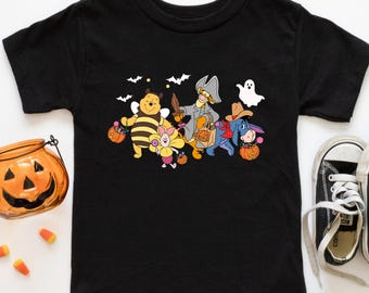 May include: Black toddler t-shirt featuring a Halloween-themed graphic of Winnie the Pooh characters dressed in costumes, including a bee, pirate, and cowboy. The design includes bats, a ghost, and pumpkins. A jack-o'-lantern and candy corn are visible.