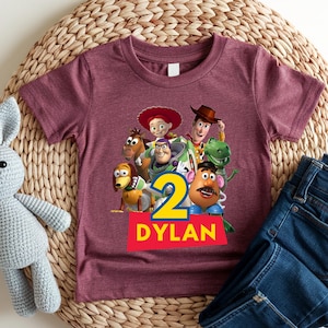 May include: Maroon short-sleeve t-shirt featuring a colorful graphic of characters from the Toy Story movie. The number "2" is displayed in yellow and blue, with the name "DYLAN" in white on a red banner.