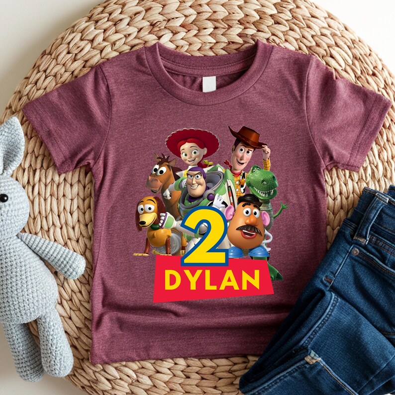 May include: Maroon short-sleeve t-shirt featuring a colorful graphic of characters from the Toy Story movie. The number "2" is displayed in yellow and blue, with the name "DYLAN" in white on a red banner.