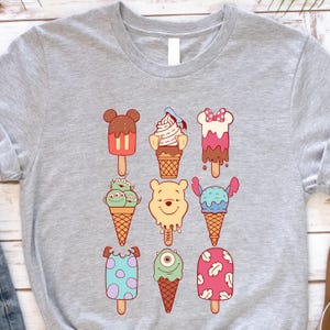 May include: A light gray t-shirt featuring colorful cartoon ice cream designs. The ice cream cones and popsicles have various character-inspired designs, including Mickey Mouse, Winnie the Pooh, and other popular characters. The shirt is made of a soft material.