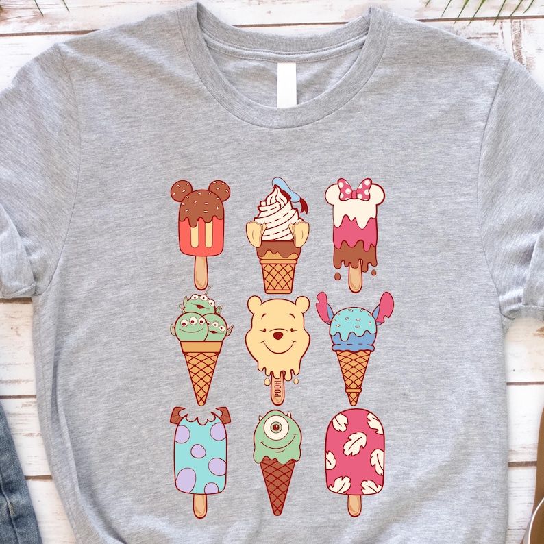 May include: A light gray t-shirt featuring colorful cartoon ice cream designs. The ice cream cones and popsicles have various character-inspired designs, including Mickey Mouse, Winnie the Pooh, and other popular characters. The shirt is made of a soft material.