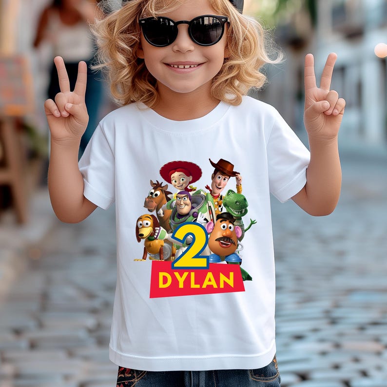 May include: White t-shirt featuring a colorful graphic of Toy Story characters, the number 2, and the name "DYLAN" in red letters. The shirt is a children's birthday theme.