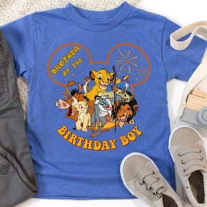 May include: A blue t-shirt featuring a graphic of characters from The Lion King, including Simba, Timon, Pumbaa, and Scar. The shirt has the text "Brother of the Birthday Boy" in orange lettering. The shirt is paired with gray pants and sneakers.