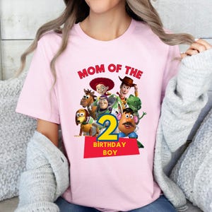 May include: Pink t-shirt featuring the phrase "MOM OF THE" in red, with a graphic of Toy Story characters and the number "2" in yellow. The text "BIRTHDAY BOY" is below the number.