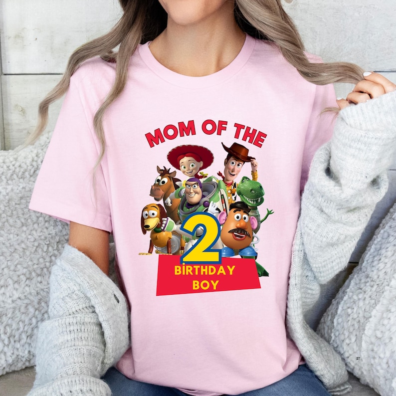 May include: Pink t-shirt featuring the phrase "MOM OF THE" in red, with a graphic of Toy Story characters and the number "2" in yellow. The text "BIRTHDAY BOY" is below the number.