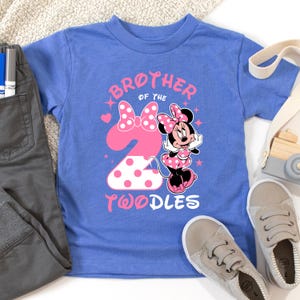 K&ouml;nnte beinhalten: Ein blaues Kleinkind-T-Shirt mit einem Minnie Mouse-Motiv. Das Shirt tr&auml;gt den Text "Brother of the 2 Twoodles" in Pink. Die Zahl 2 ist rosa mit wei&szlig;en Punkten. Eine graue Hose und Schuhe liegen neben dem Shirt.