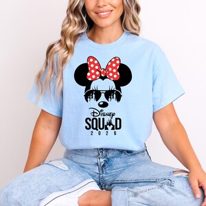 May include: Light blue t-shirt featuring a Minnie Mouse graphic with sunglasses and a red polka dot bow. The text "Disney Squad 2026" is printed below the graphic. The shirt is a casual style.