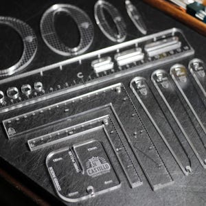 May include: A collection of clear acrylic rulers and templates, including circles, straight edges, and curved shapes. The rulers are marked with inch measurements and the brand name "Castillo". The tools are arranged on a dark surface.