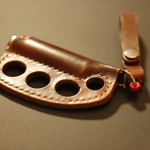 May include: A brown leather key chain with a brass ring and a red string. The key chain has a leather loop with four holes for fingers.