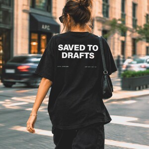 May include: A black short-sleeve t-shirt featuring the white text SAVED TO DRAFTS printed on the back. Below the main text, smaller white text reads STATUS: UNPUBLISHED and LAST EDIT: 02:37 AM. The shirt has a loose fit and a crew neckline.