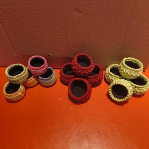 Puede incluir: Una colección de pequeños cuencos de ganchillo en varios colores, incluyendo rojo, verde, amarillo y rosa. Cada cuenco tiene un interior oscuro y un exterior texturizado. Los cuencos están dispuestos en pequeños grupos sobre una superficie naranja.