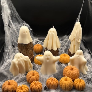 May include: A Halloween-themed display featuring ivory-colored ghost candles and small orange pumpkin decorations. The candles are in various shapes, some perched on wooden bases. The scene is set against a backdrop of faux spiderwebs and black accents.