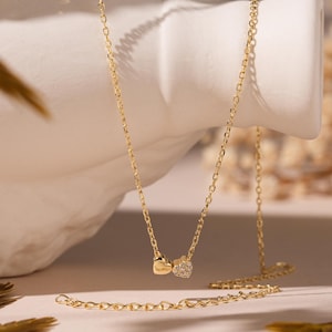 May include: A delicate gold necklace with two pendant charms. One charm is a small heart, and the other is a square shape with small, sparkling stones. The necklace is displayed on a white, textured surface with dried foliage.