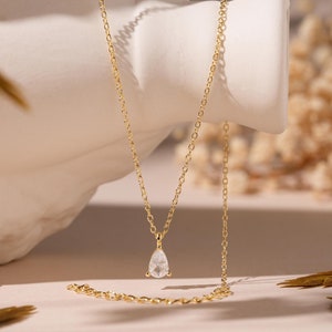 May include: A delicate gold chain necklace with a pear-shaped clear gemstone pendant. The necklace is displayed on a white surface with dried flowers in the background.