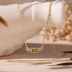 May include: A gold chain necklace with a small rectangular pendant featuring a clear stone. The pendant is accented with smaller stones on either side.