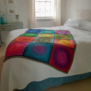 May include: A white bed with a colorful crocheted throw blanket. The blanket is made up of 9 squares, each with a different mandala design. The colors of the blanket are red, orange, yellow, green, blue, purple, and pink.
