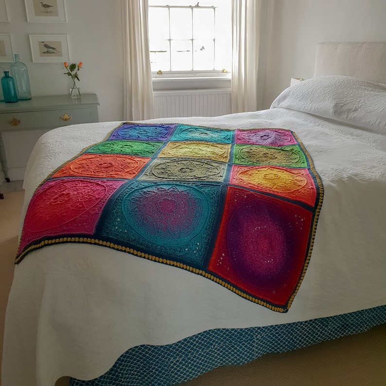 May include: A white bed with a colorful crocheted throw blanket. The blanket is made up of 9 squares, each with a different mandala design. The colors of the blanket are red, orange, yellow, green, blue, purple, and pink.