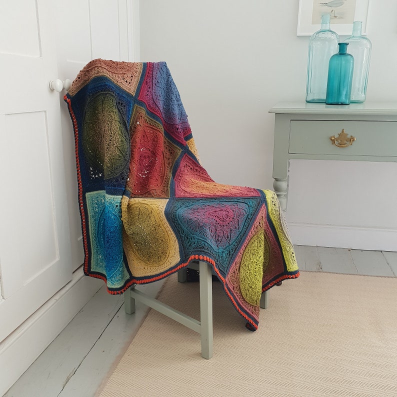 May include: A colorful crocheted throw blanket with a geometric pattern in shades of blue, green, red, and brown. The blanket is draped over a wooden chair with a light green cushion.