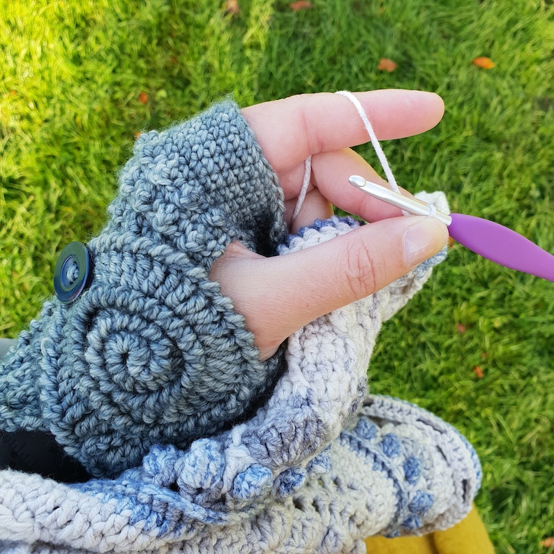 Op de afbeelding: Gehaakte vingerloze handschoenen in grijstinten en blauw, met een spiraalpatroon en knoopdetail. Een haaknaald en wit garen worden gebruikt om het werk voort te zetten. De achtergrond is wazig groen gras.