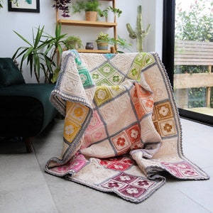 Puede incluir: Una manta de ganchillo con un patrón floral colorido. La manta está hecha de hilo blanco con cuadrados de diferentes colores, incluyendo rosa, verde, naranja y morado. La manta está doblada y drapeada sobre una silla.