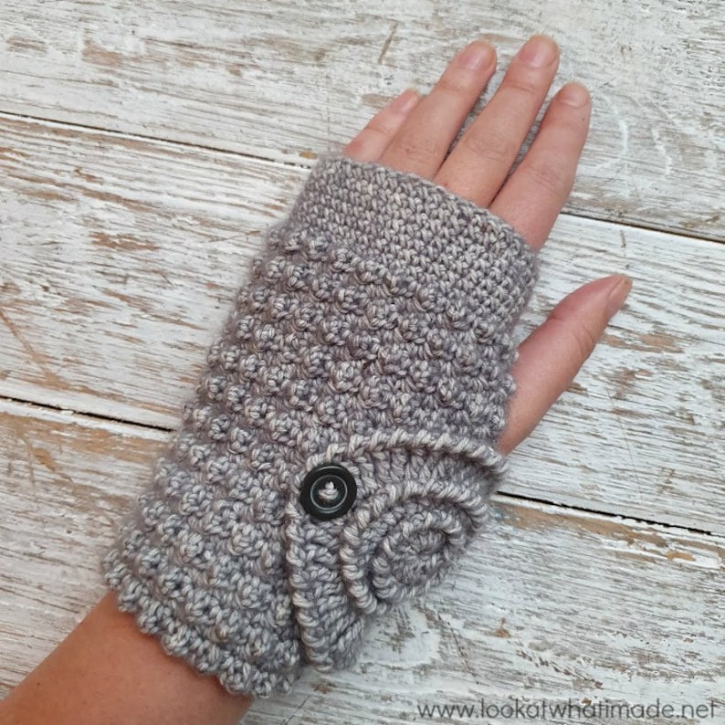 Op de afbeelding: Een grijze gehaakte vingerloze handschoen met een textuurpatroon en een knoopsluiting. De handschoen is gemaakt van zacht, getextureerd garen en is ontworpen om de handen warm te houden terwijl de vingers vrij blijven.