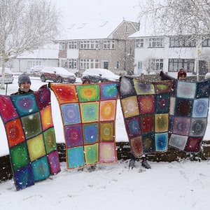 May include: Four people stand in the snow holding up colorful crocheted blankets. The blankets are made of squares with a circular pattern in the center of each square. The blankets are in shades of red, orange, yellow, green, blue, purple, and brown.