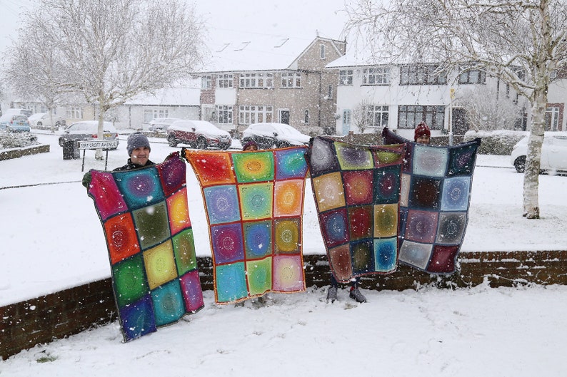 May include: Four people stand in the snow holding up colorful crocheted blankets. The blankets are made of squares with a circular pattern in the center of each square. The blankets are in shades of red, orange, yellow, green, blue, purple, and brown.