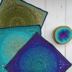 May include: Three crocheted square blankets in shades of green, blue, and purple. The blankets are layered on top of each other, with the largest blanket on the bottom and the smallest blanket on top. The blankets are made with a variety of stitches and patterns.