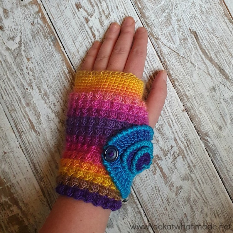 Op de afbeelding: Een handgemaakte, vingerloze gehaakte handschoen in een levendige regenboog van kleuren, waaronder geel, oranje, roze, paars en bruin. Een blauw, decoratief accent met een knoopsluiting is aan de pols bevestigd. De handschoen wordt op een hand getoond.